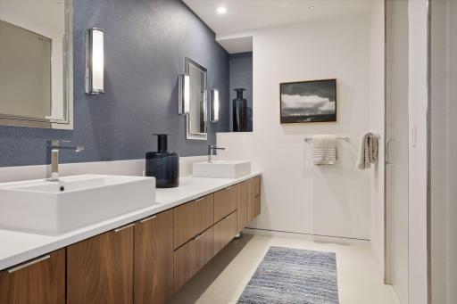 Private en suite bath for the main level bedroom featuring a double vanity, walnut cabinetry, tile floors, medicine cabinets, and etched glass doors to the walk-in shower and separate water closet.