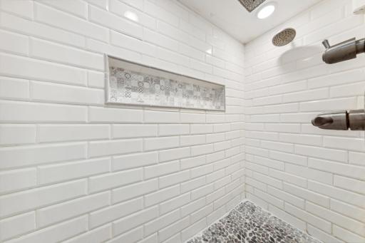 View of lower-level shower, with subway tile walls, pebbled, mosaic tiled floor, and large niche.