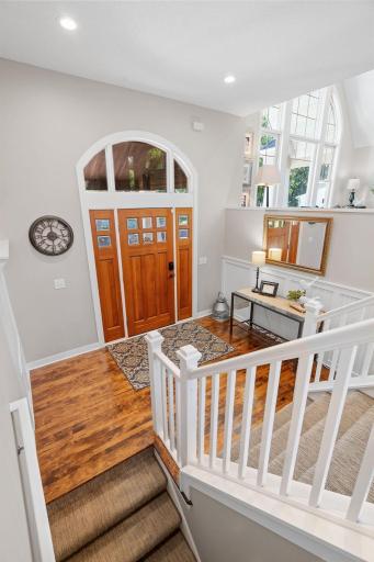 Foyer features maple hardwood flooring finished in a warm walnut tone, enameled wainscoting w/ hooks, craftsman solid wood entry door w/ decorative arched top window, and open, wide staircase w/ enameled railings
