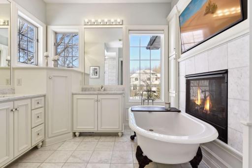 Spa-like primary bath with marble floors, dual vanities, footed tub with dual-sided fireplace, & tons of sunlight!