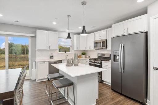 (Photo of decorated model, actual home's finishes will vary) Designer kitchen with white cabinetry, quartz countertops, slate-finish appliances, gas stove, backsplash, deep sink, center island, and pantry closet.