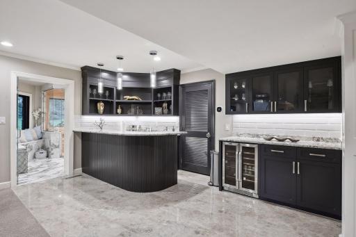 To the left lies a well-appointed bar, finished in sleek black with granite countertops, beverage fridge, microwave, and sink. The tile floor and subway tile backsplash add a touch of sophistication to the space.