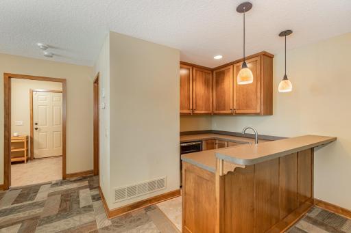 The wet bar has: a recessed light, 2 pendant lights, custom maple cabinets, stainless steel sink, a wine fridge & ceramic tile floor. Just behind the wet bar is the 3rd bedroom. You can also see the entrance into: the LL mudroom area & full bathroom.