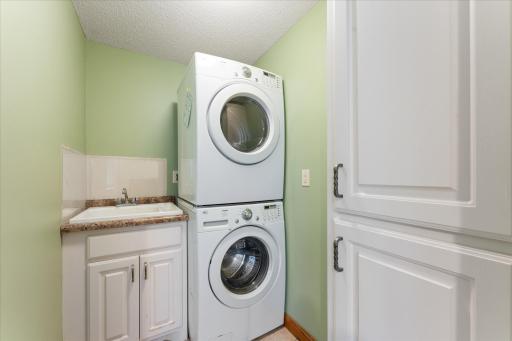 Lower-level laundry room includes stacked washing machine and dryer next to sink with cabinet and linen closet.