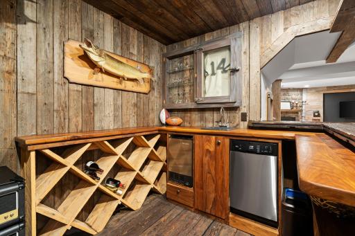 Full bar made from rustic barn wood features exotic wood countertops with quartz sink, wine bottle storage, display cabinet, dishwasher, ice maker, an area for a beverage refrigerator and raised countertop.