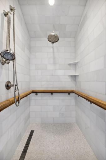 The curbless marble-tiled shower blends elegance and accessibility, featuring dual shower heads and a reinforced handrail for added comfort, safety, and seamless style.