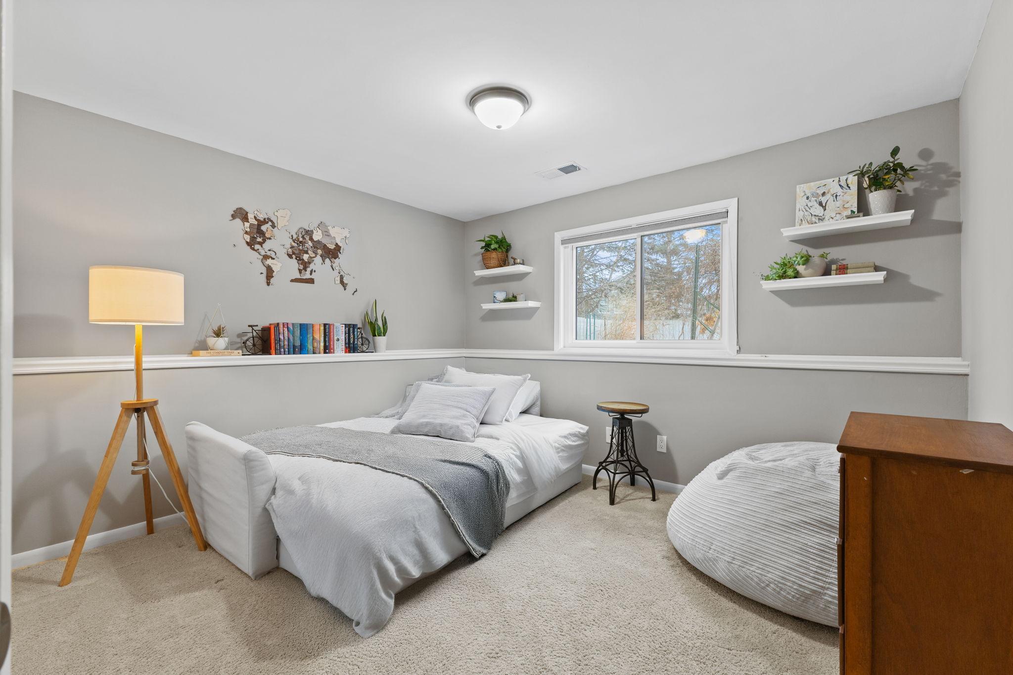This lower-level bedroom boasts generous space and large windows that overlook the lush backyard. There is a large closet to the left of the lamp.