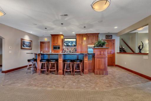 The wet bar in the lower-level family room is designed for entertaining in style. It features rich custom cherry wood cabinetry, a spacious countertop with ample bar seating, and modern pendant lighting that adds a warm ambiance.