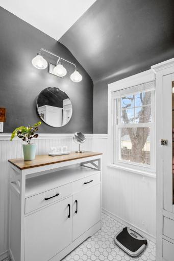 A charming and light-filled vanity nook offers a peaceful spot to get ready, complete with a round mirror, updated lighting, and a beautiful blend of white wainscoting and bold wall color.