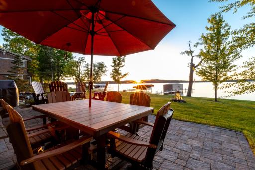 Lakeside dining patio with firepit, umbrella seating, and panoramic sunset views over the water.