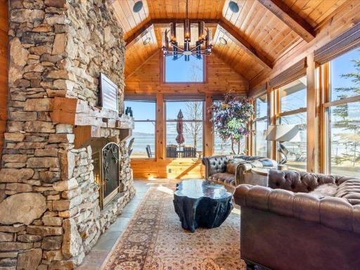 Sunlit lakeside living room with vaulted wood ceilings, floor-to-ceiling windows, and grand stone fireplace.