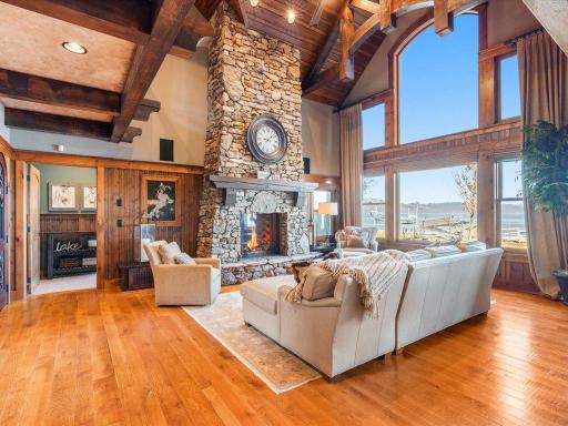 Elegant great room with stone fireplace, exposed beams, and panoramic lake views through floor-to-ceiling windows.