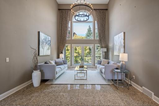 The formal living room impresses with an 18-foot ceiling, a stylish chandelier, marble tile floors, and glass French doors that lead to the outdoor deck, creating a bright and elegant atmosphere.