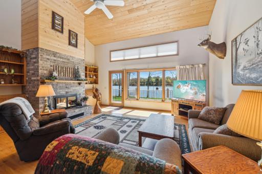 Bright and inviting living area with tongue-and-groove ceiling, full-height fireplace, and panoramic lake views through large windows.