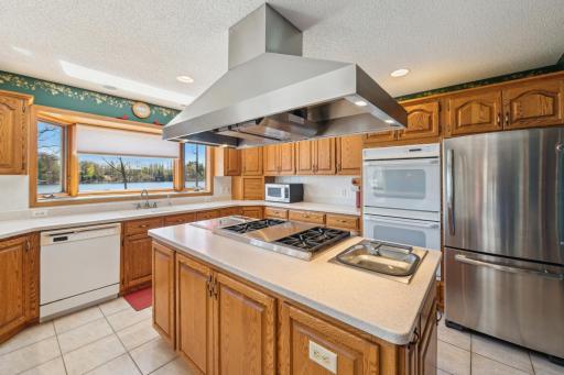 Chefâs kitchen with stainless steel hood, large island cooktop, double ovens, and picture window showcasing Long Lake.