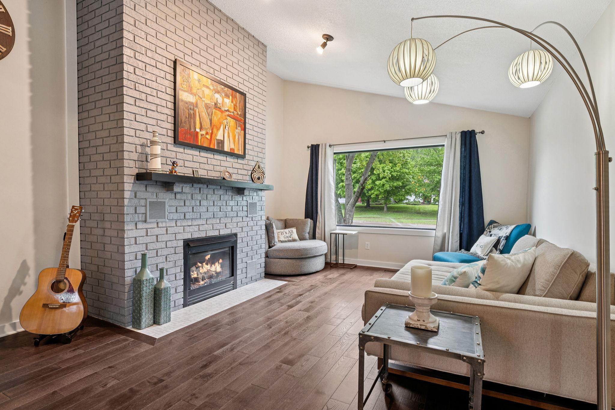 Warm and welcoming living room with rich hardwood floors, a stunning painted brick gas fireplace, and large picture window filling the space with natural light.