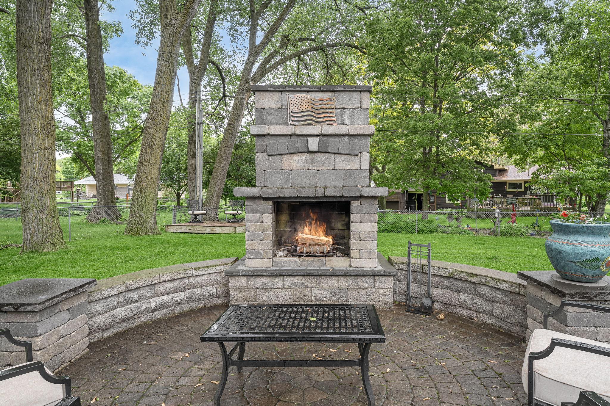 Stunning stone outdoor fireplace creates a cozy focal point for your backyard. Surrounded by curved seating and mature trees, it's perfect for hosting or unwinding by the fire.