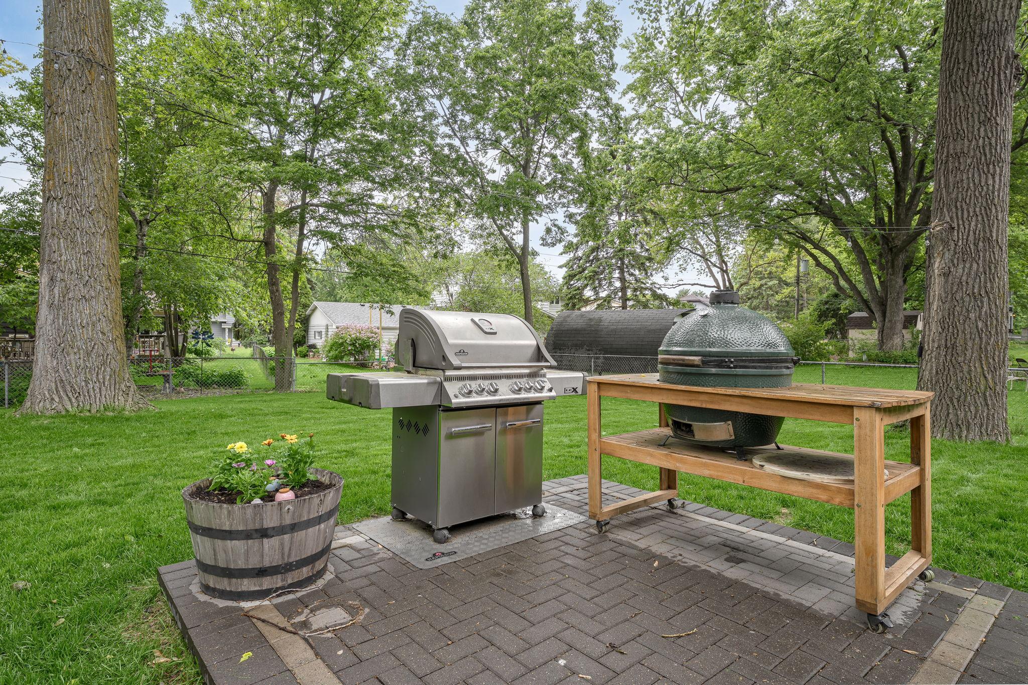 Backyard grilling station with both a gas grill and ceramic smoker, set on a paved pad surrounded by green space, ideal for cookouts and summer gatherings.