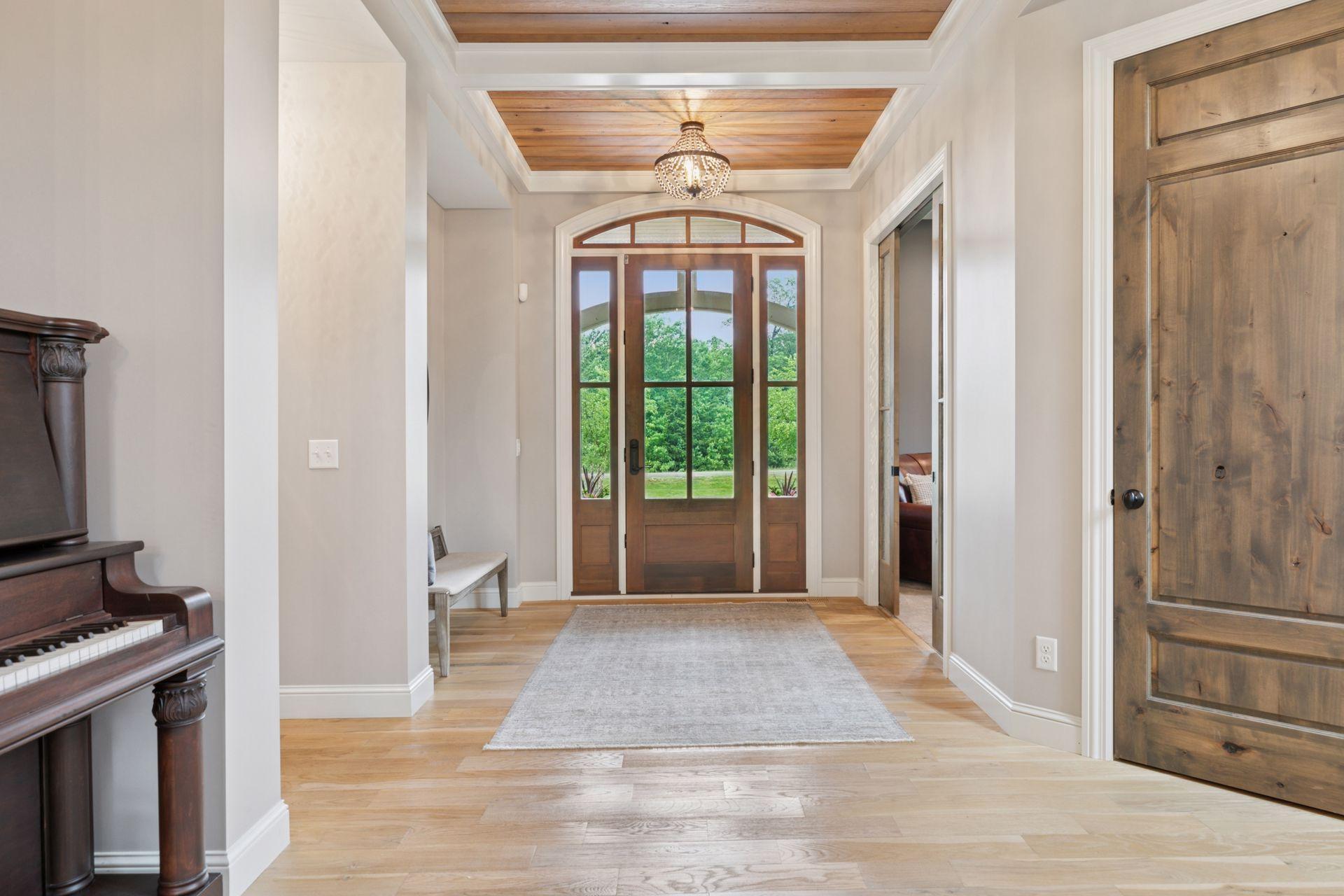 Inviting front entry showcases warm wood accents, coffered ceiling detail, and natural light streaming through full-view glass doors with scenic views of the lush front yard.