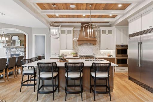 Custom chefâs kitchen featuring a large center island with seating, built-in double ovens, statement hood, and wood-inlaid coffered ceiling.