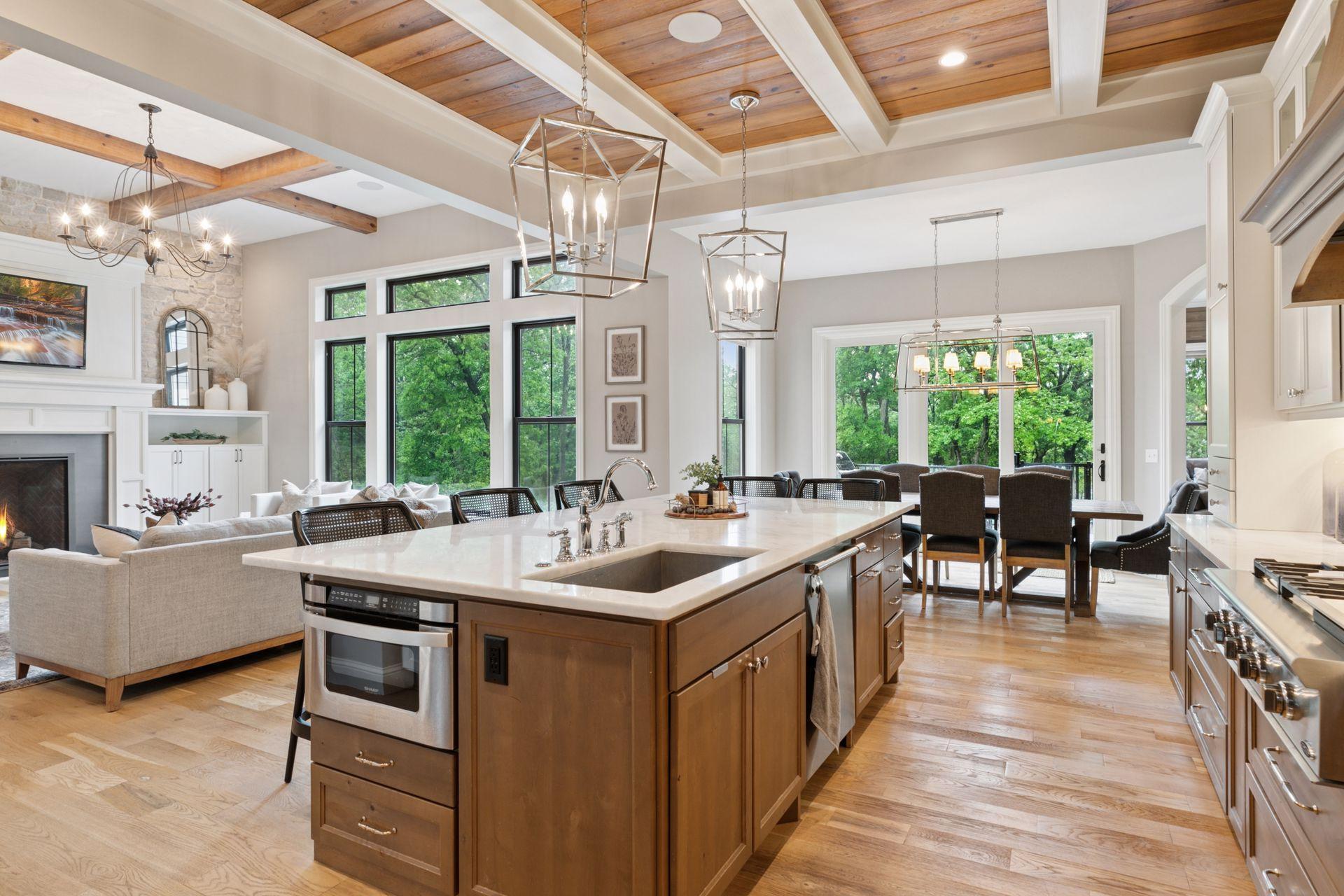 Expansive kitchen island anchors the space, offering views of the great room, dining area, and wooded landscape beyond tall windows.