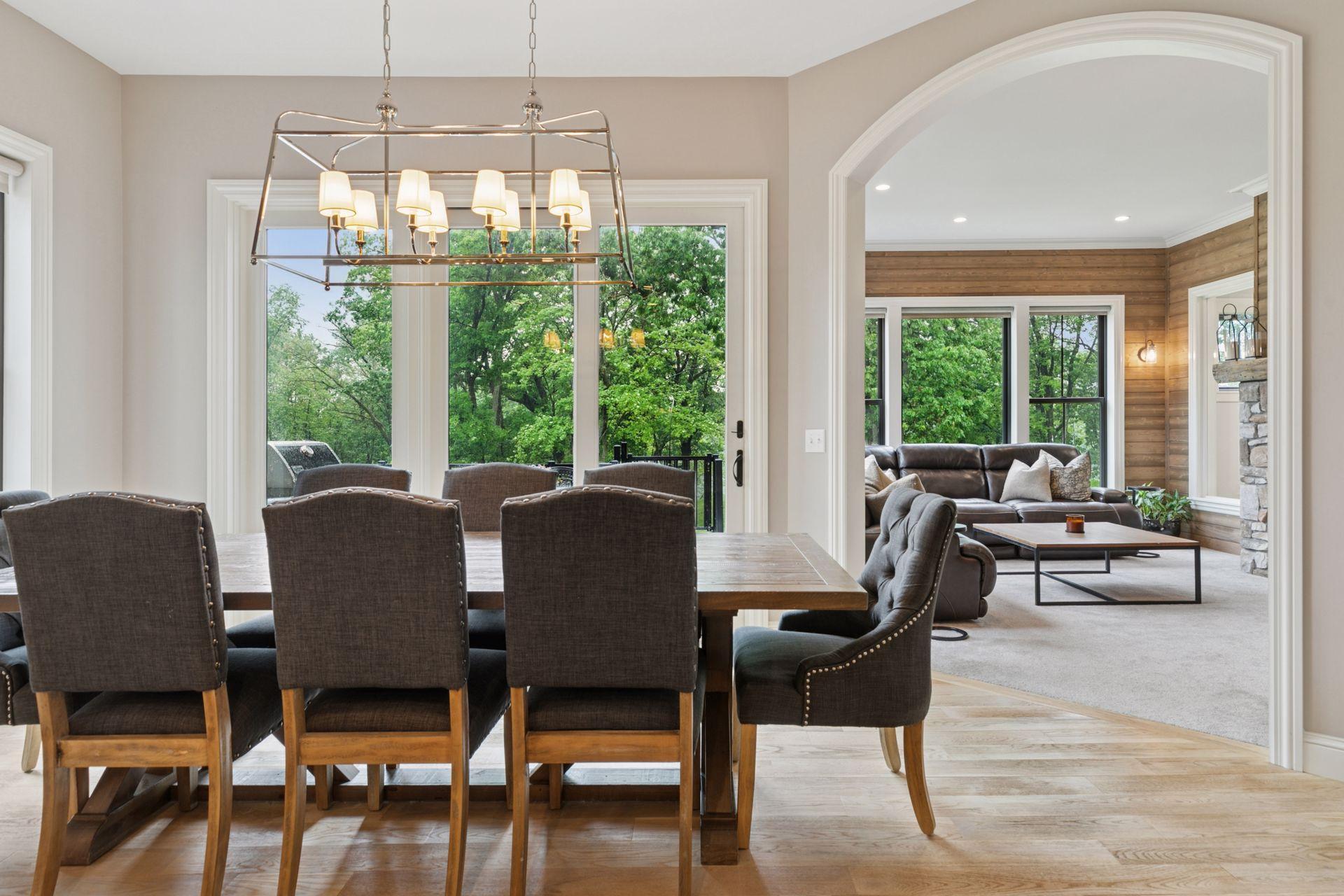Elegant formal dining room, bathed in natural light from expansive windows and glass doors. Beautiful archway leads to a cozy sunroom with stone fireplace and wood-paneled walls.