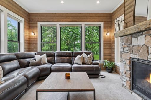 Charming sunroom with wraparound windows offering tranquil views of the wooded backyard. Features include wood accent walls, neutral carpeting, and a stunning floor-to-ceiling stone fireplace with rustic mantle.