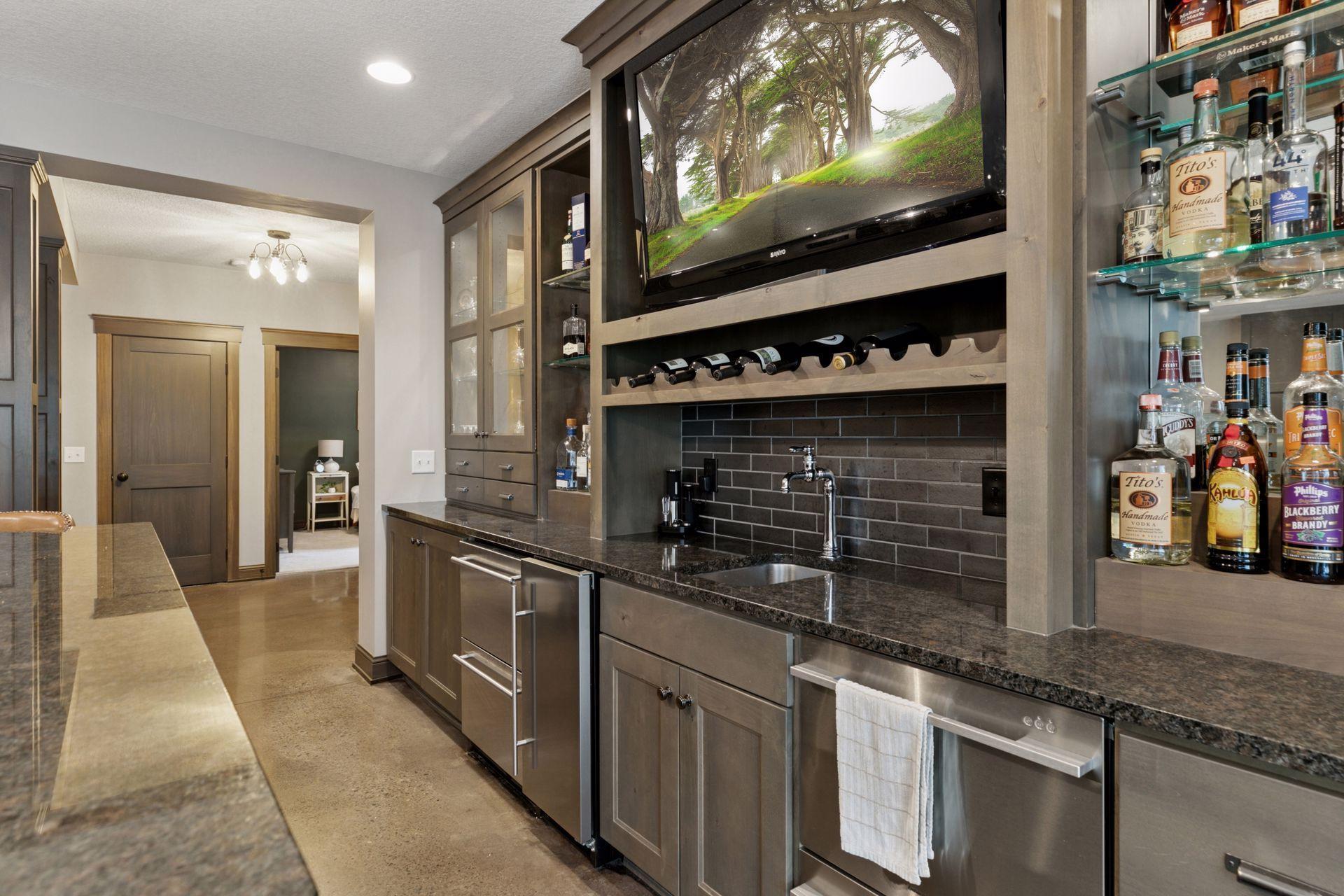 Stylish wet bar features rich wood cabinetry, a sleek dark countertop, built-in wine rack, under-counter refrigeration, and a wall-mounted TV. Glass shelves display a curated liquor collection, making this the perfect spot for entertaining.