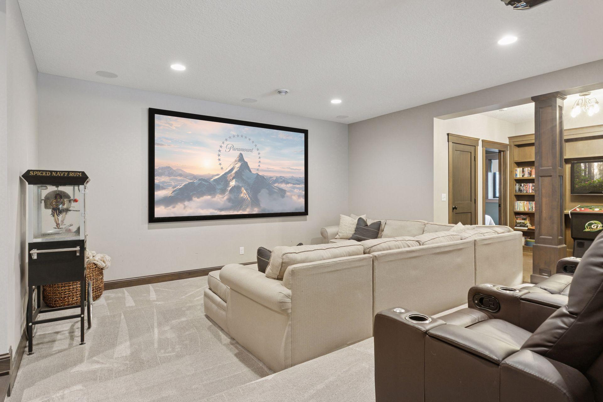 Spacious and inviting home theater featuring a plush sectional, cozy ottomans, and an oversized wall-mounted screen—perfect for movie marathons or game day. Soft carpeting and warm neutral finishes create a relaxed, immersive atmosphere.
