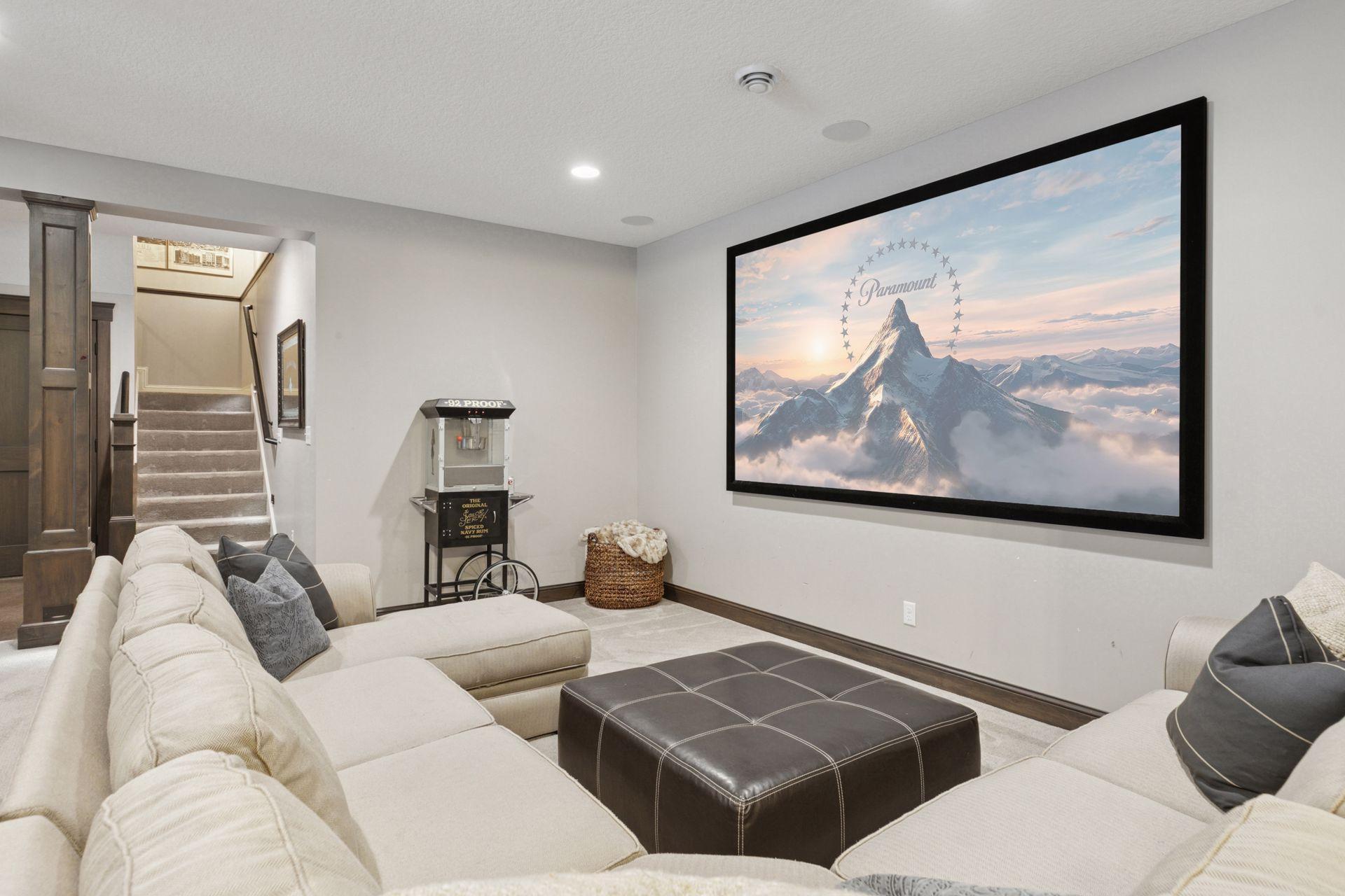 Cozy home theater setup with a large sectional, ottoman seating, and a massive wall-mounted screen ideal for movie nights. Neutral tones and soft carpeting enhance the comfort and ambiance of this entertainment space.
