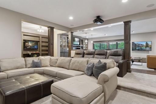 Expansive lower-level entertainment area with tiered theater seating, arcade nook, and an open view to the bar and lounge. Recessed lighting and a ceiling-mounted projector complete the perfect setup for hosting family and friends.