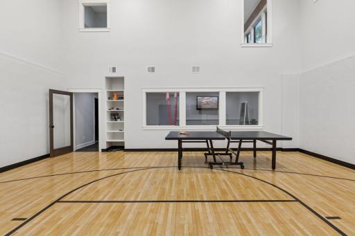 Sport court with polished hardwood floors, soaring ceilings, ping pong table, and built-in shelvingâseamlessly connected to the fitness room for active living.