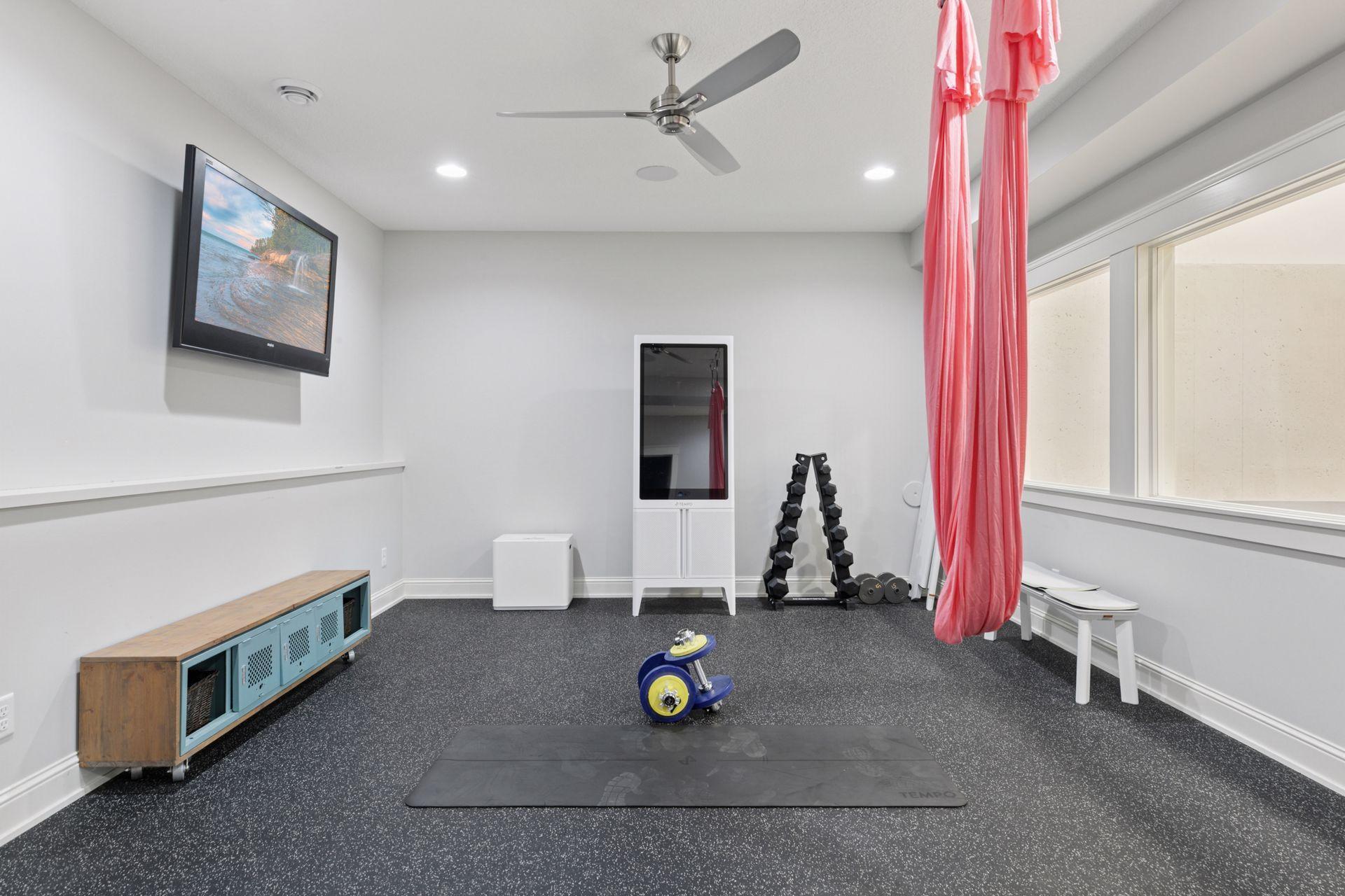 Bright, modern fitness room with rubber flooring, wall-mounted TV, ceiling fan, and aerial yoga setup—ideal for strength training, stretching, and cardio.