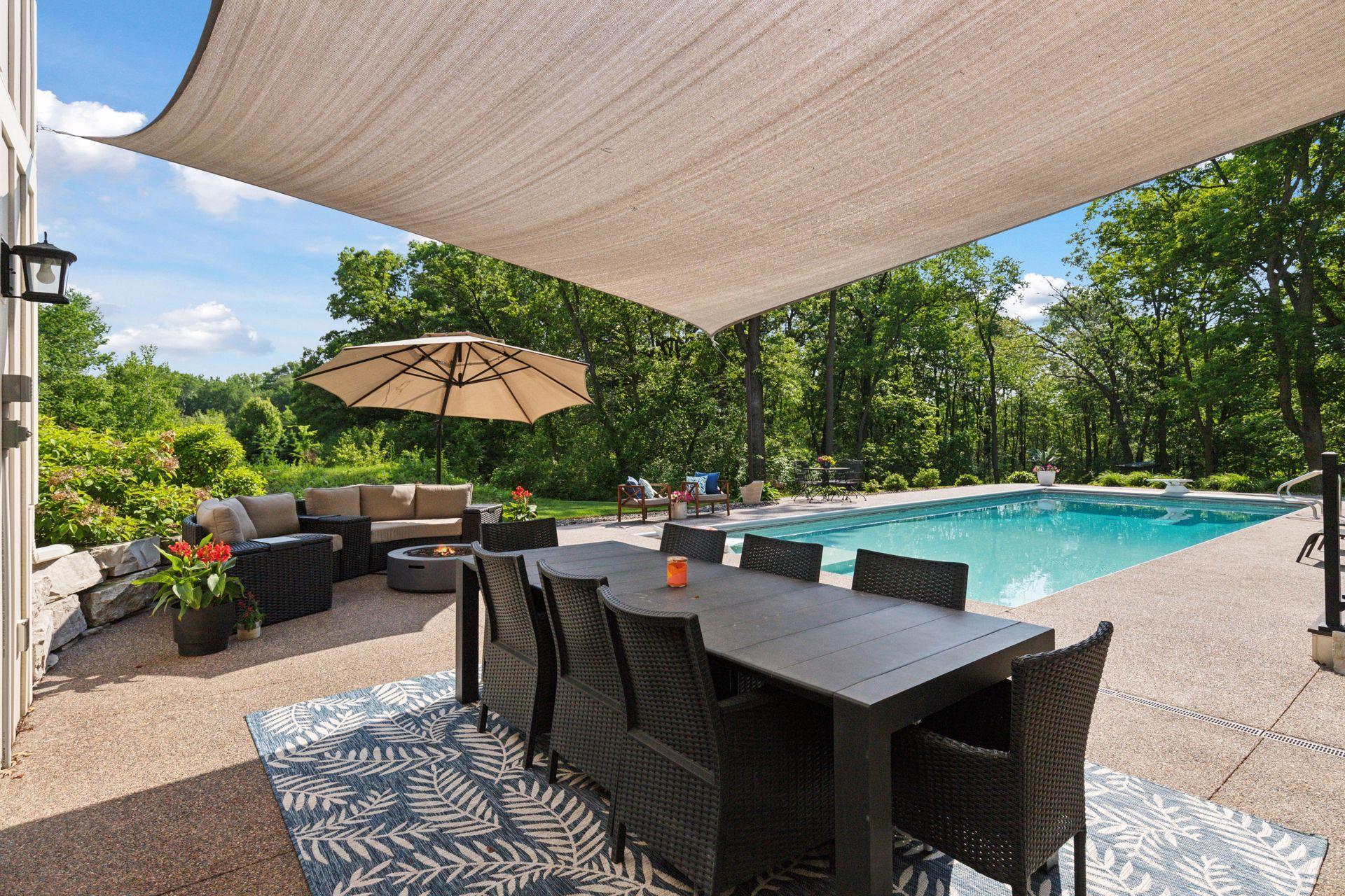 Shaded poolside dining area with large sun sail, firepit, and lounge seating. Perfect for outdoor meals and relaxing in comfort with scenic wooded views.