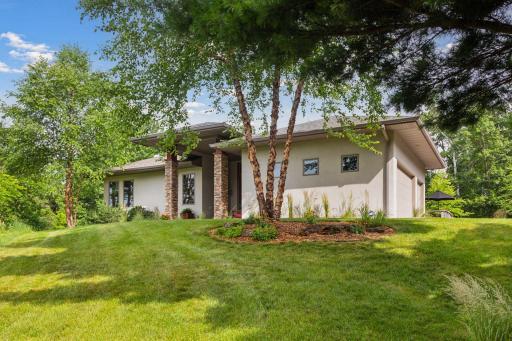 The front of the home makes a bold first impression with its modern one-level design, crisp stucco exterior, and striking stone pillars—all set against a lush, tree-filled backdrop.
