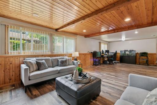Warm and welcoming family room featuring a cozy brick fireplace, wood-paneled ceiling and walls, and large windows that fill the space with natural light â perfect for relaxing or entertaining.