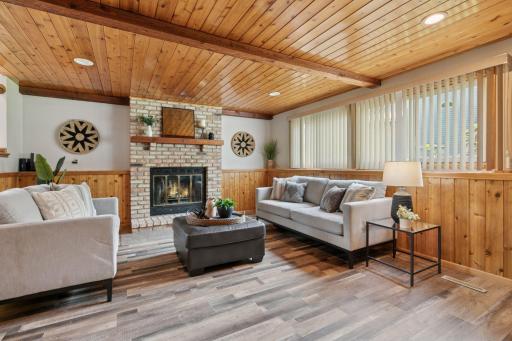 Warm and welcoming family room featuring a cozy brick fireplace, wood-paneled ceiling and walls, and large windows that fill the space with natural light â perfect for relaxing or entertaining.