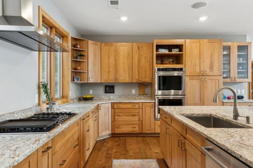 This chef-inspired kitchen features rich wood cabinetry, double ovens, granite countertops, and a gas cooktop with sleek stainless vent hood. Thoughtful details like open corner shelving and glass-front cabinets add both charm and function.