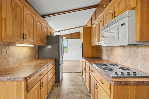 This view highlights the cooktop, newer wall oven, newer fridge, and a microwave. Beautiful hickory cabinets offer endless storage, while the seamless transition directly into the dining room makes everyday entertaining a breeze.