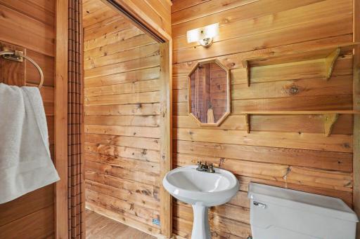 Convenient lower-level half-bath in the sunroom features LVT flooring, warm knotty pine woodwork, and an elegant white pedestal sink. Ideal for guests and quick indoor/outdoor access.