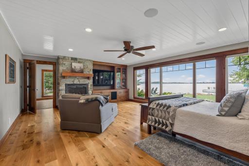 The primary suite is a private retreat with wood flooring, a wood accent wall, large picture windows overlooking the lake, and a stone-surround gas fireplace.