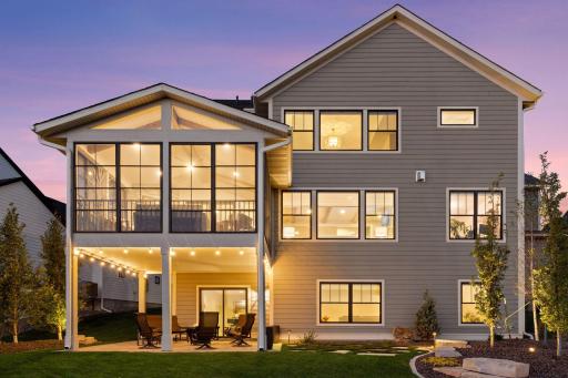 The back of the home glows warmly at twilight, showcasing the inviting three-seasons porch above and a spacious patio below, perfectly designed for outdoor seating and evening gatherings