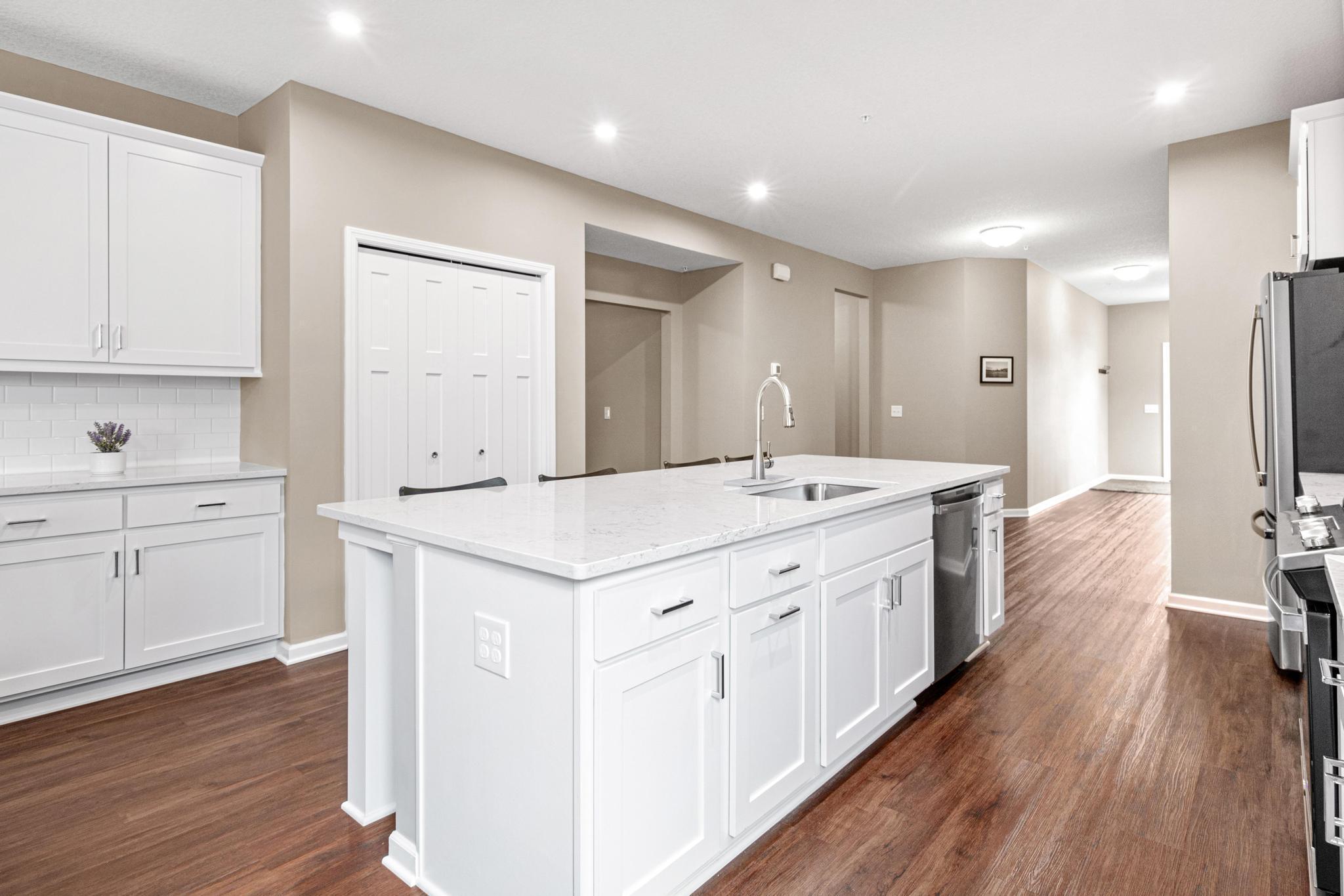 Gourmet kitchen view looking toward the entry and stairs, highlighting the seamless main-level flow. This perspective showcases the coffee/bar area, double-door pantry, and wide hallway leading to the upper-level loft and guest suite.