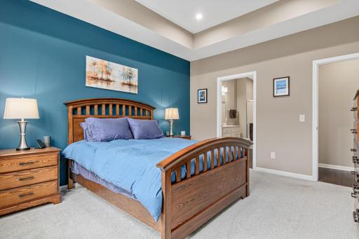 Primary suite view toward the private ensuite entrance. Features a tray ceiling, stylish accent wall, and plush carpet. This oversized main-floor retreat offers a seamless flow and abundant space for relaxation in a modern layout.
