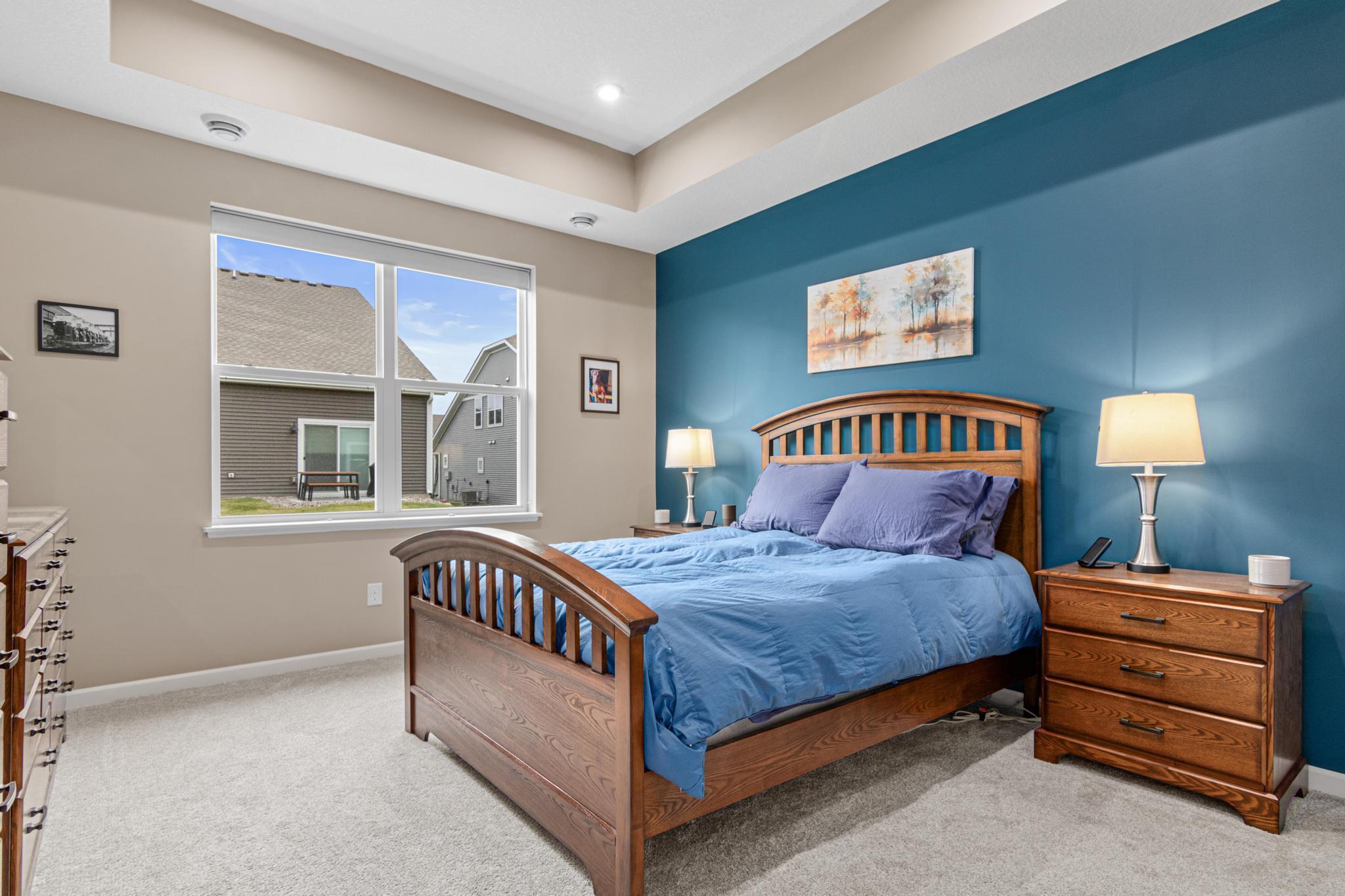 Oversized main-floor primary suite with a stylish accent wall, tray ceiling, and plush carpet. A large window floods the room with light, offering a spacious and private retreat within this modern one-level living floorplan.