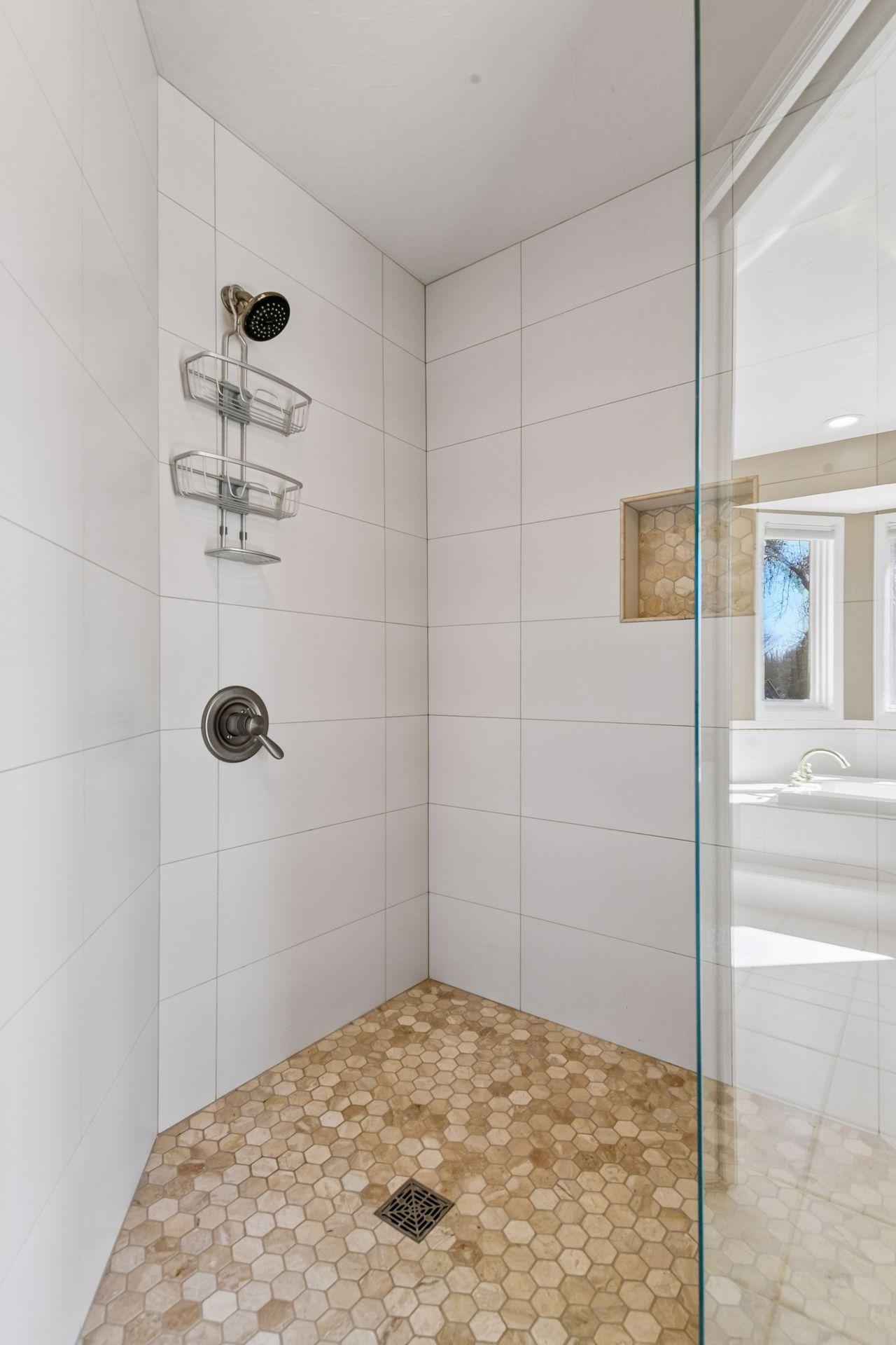 The walk-in shower features sleek floor-to-ceiling tiling and a beautiful hexagonal stone floor. A custom recessed niche and frameless glass door add a refined, spa-like touch!