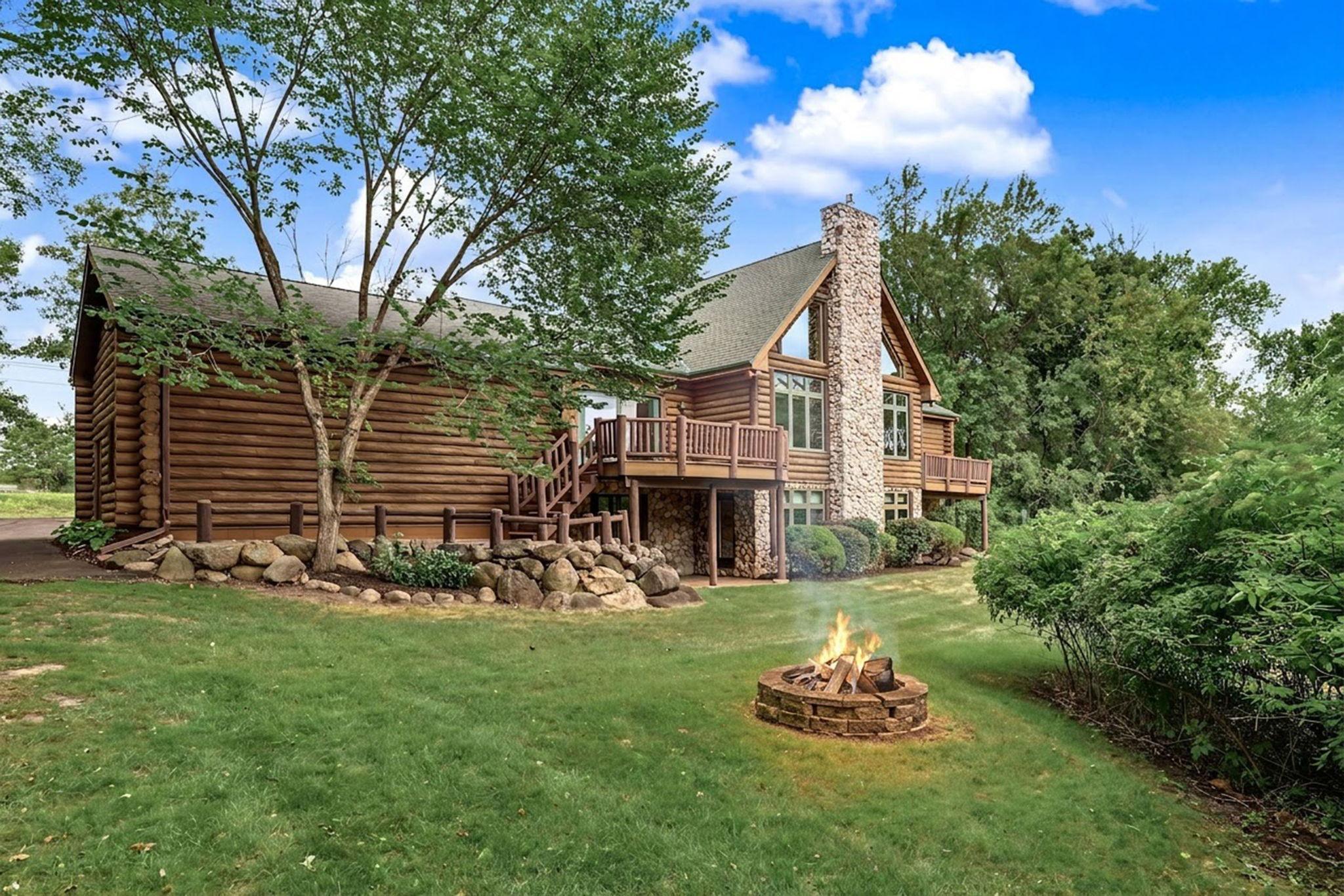 Rear exterior photo of home showing log/stone exterior, 2 decks, paver patio, dramatic stone chimney and firepit.
