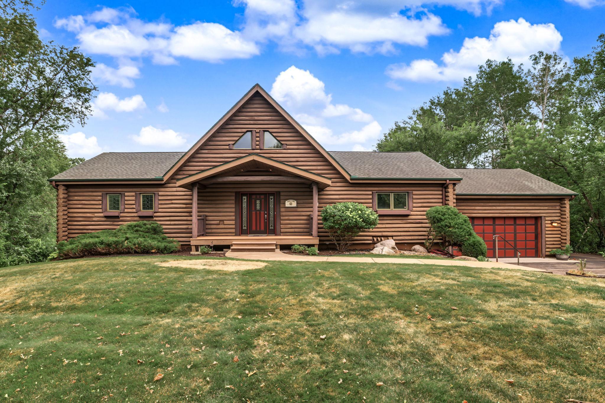 Classic log home nestled on 1.47 private acres, featuring a welcoming covered front porch, inviting entry, attached 2-car garage, and timeless Northwoods architecture surrounded by mature trees.