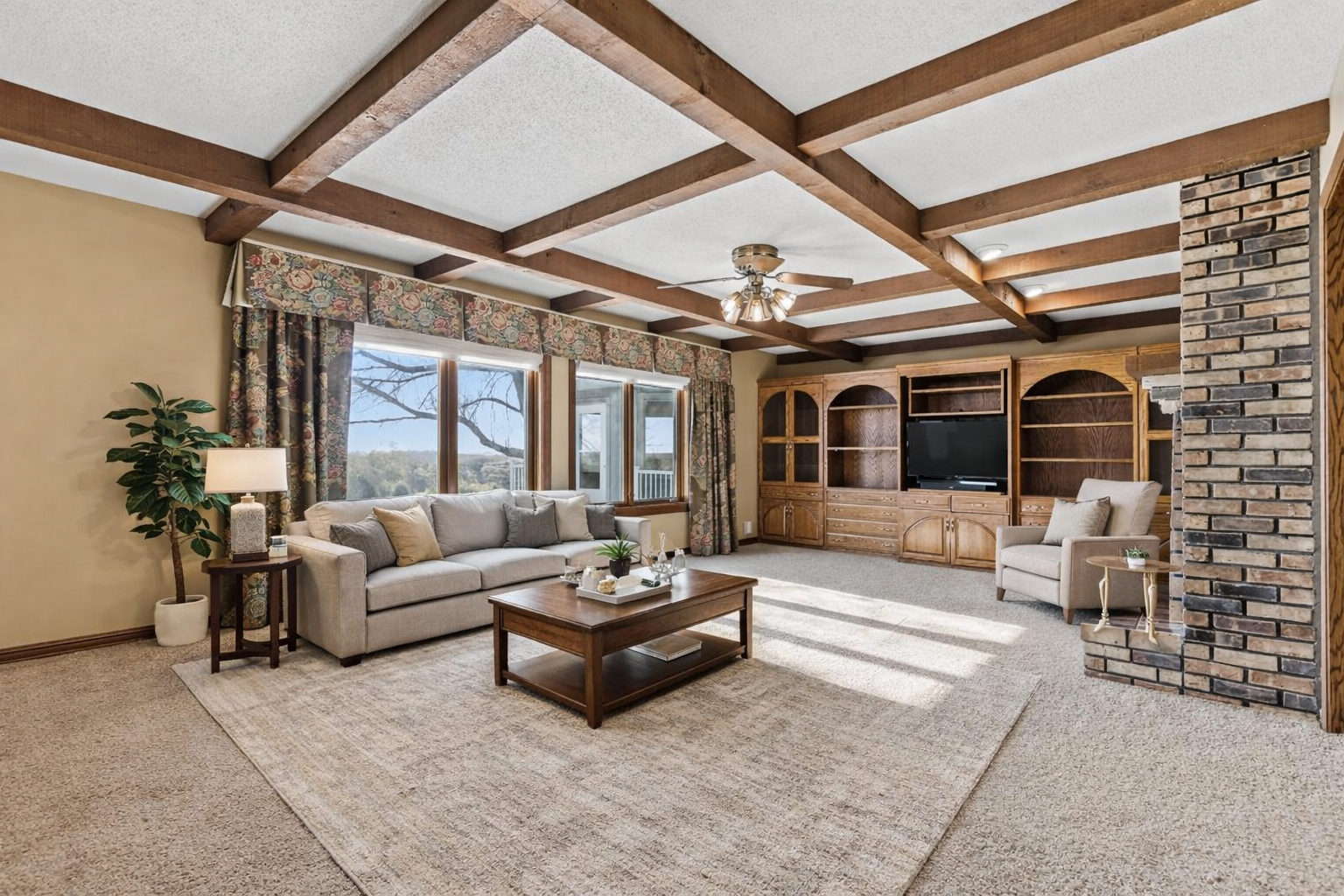 Bright and inviting living room featuring exposed wood beams, floor-to-ceiling windows, cozy fireplace, and custom built-ins—an ideal gathering space with timeless character. virtually staged