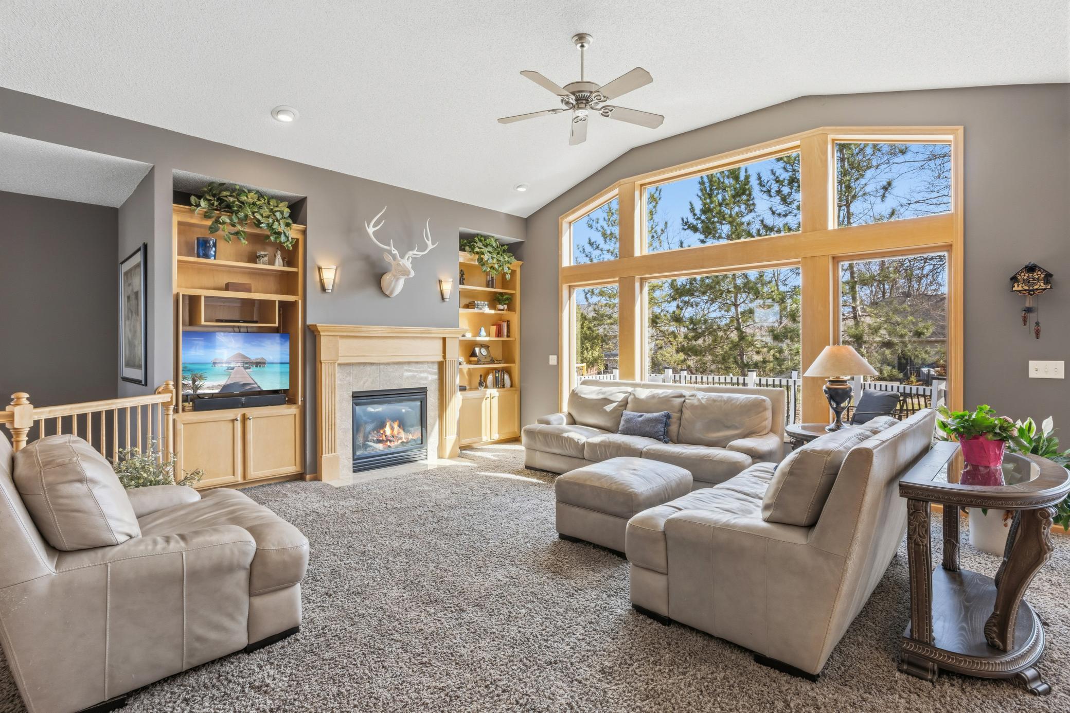 The living room is a stunning centerpiece with a soaring vaulted ceiling and floor-to-ceiling windows that flood the space with natural light. Maple built-ins add warmth and style, while the gas fireplace provides a cozy focal point.